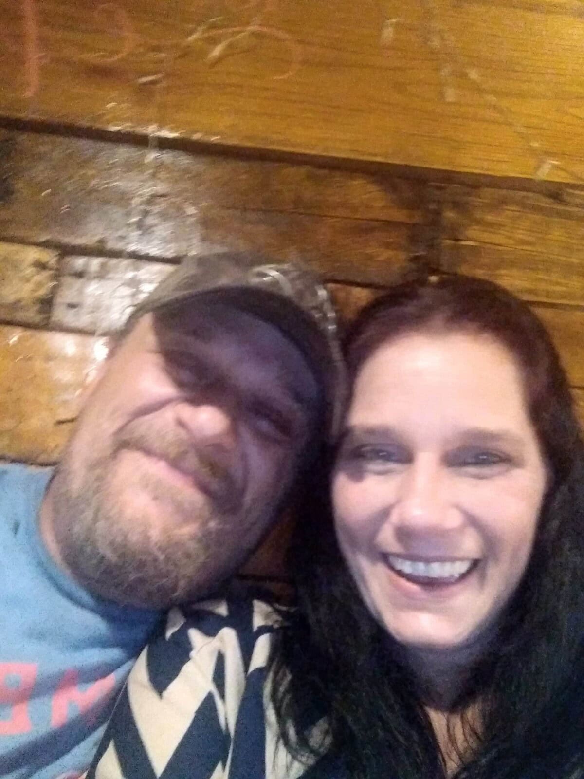 A man wearing a cap and a woman with dark hair are sitting close together, smiling at the camera in front of a wooden wall.