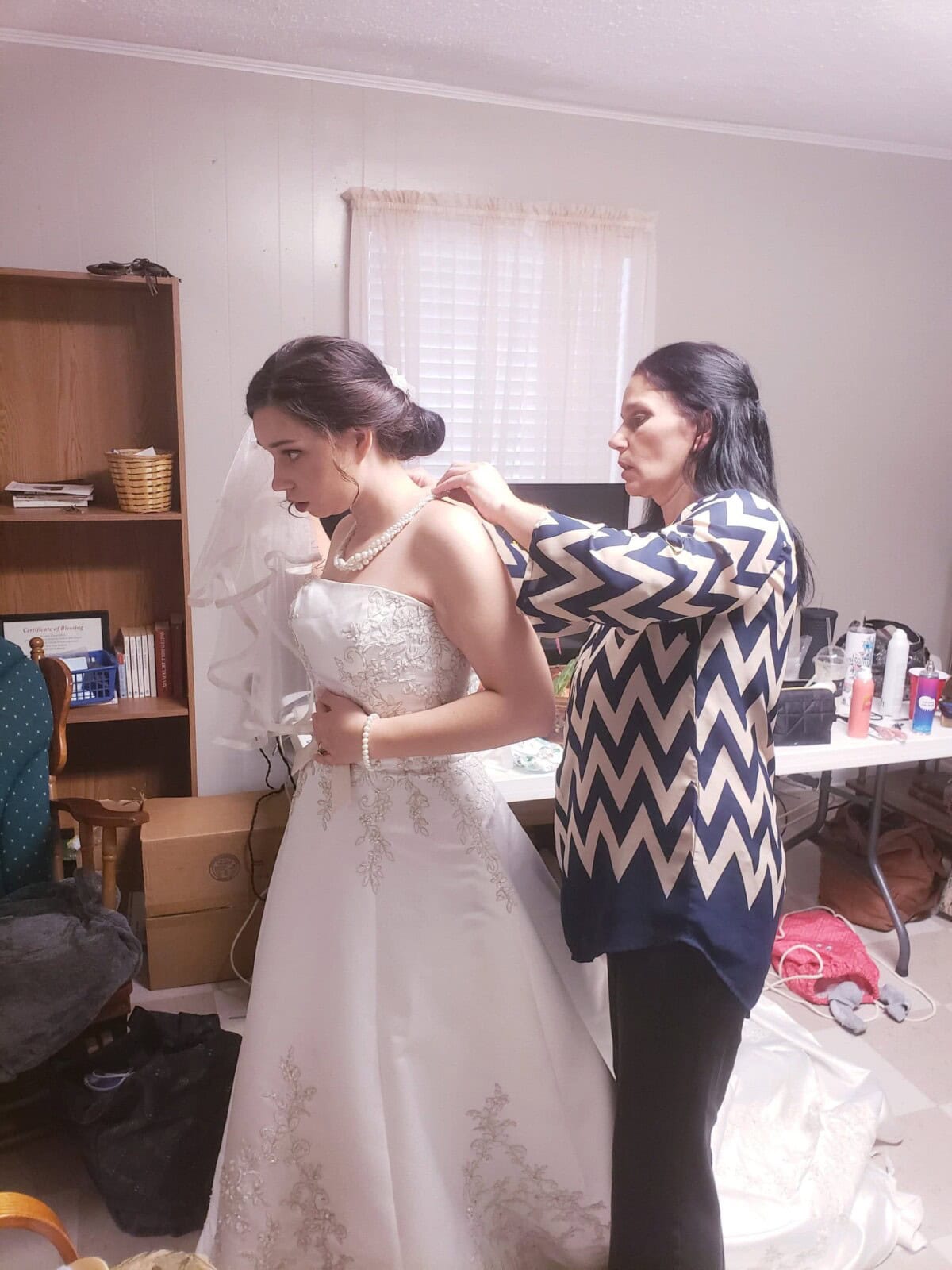 A woman assists a bride in a white wedding dress with her necklace in a cluttered room.