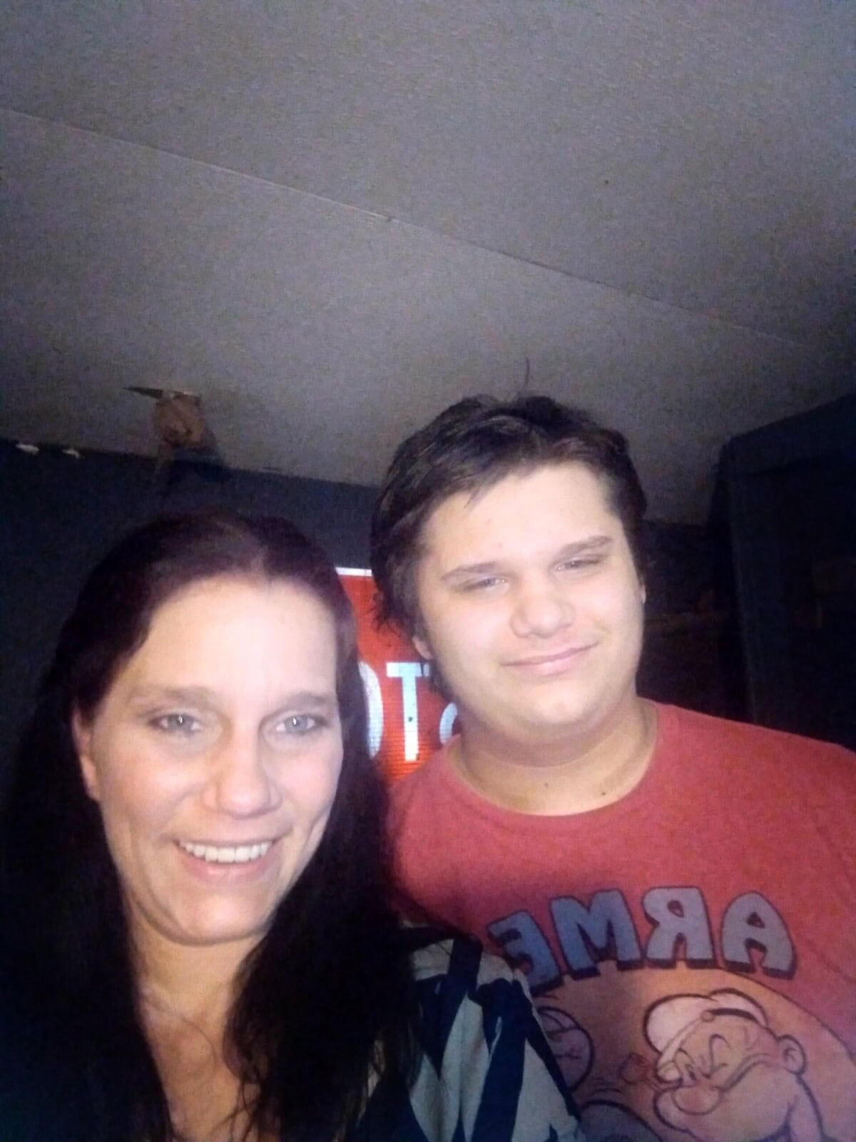 A woman and a teenage boy stand indoors, smiling at the camera. The boy is wearing a red Popeye T-shirt. The background is dimly lit.