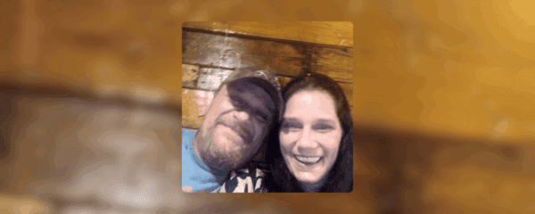 A man wearing a cap and a woman with long dark hair smile while sitting closely together against a wooden wall.