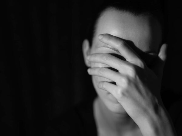 A person covers their face with one hand, partially obscuring their features, in a low-light, black-and-white setting.
