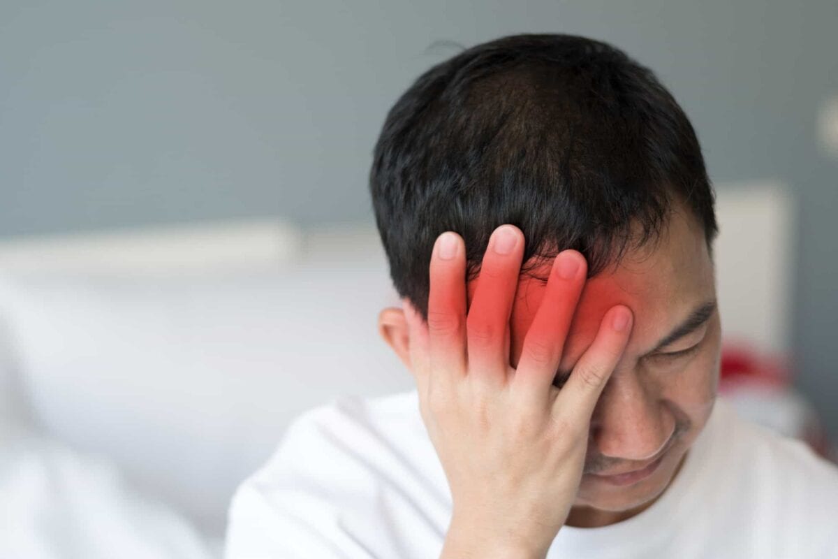 A man sitting on a bed holds his head with one hand, showing a red area on his forehead, indicating pain or headache.