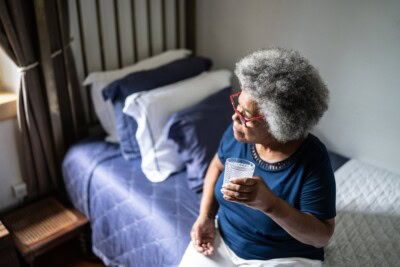 An older woman with gray hair and red glasses sits on the edge of a bed, holding a glass of water and looking out a window.