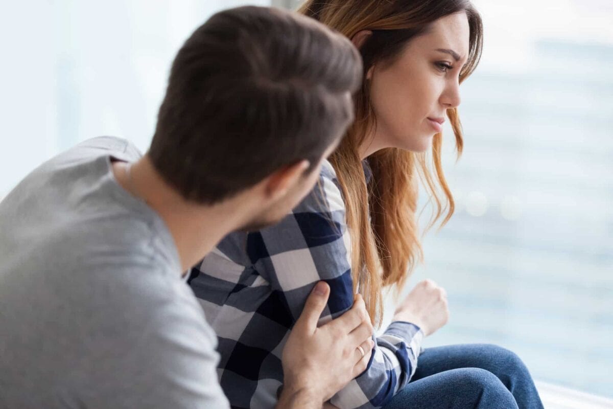 A man gently holds a woman’s arm while she looks away with a sad expression, both sitting indoors near a window.