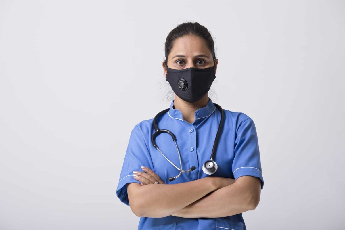 A healthcare worker wearing blue scrubs, a stethoscope, and a black face mask stands with arms crossed against a plain white background.