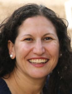 A woman with long, curly dark hair and pearl earrings smiling at the camera in an outdoor setting.