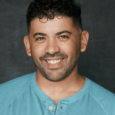 Man with short curly hair and trimmed beard smiling, wearing a light blue shirt against a dark background.