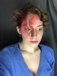 Person with curly hair wearing a blue top, with intricate red and gold face paint covering one side of their face, standing against a dark background.
