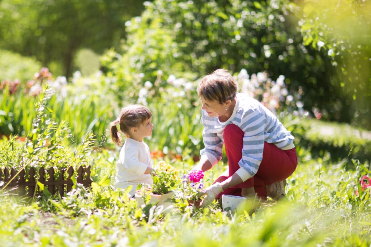 An older woman and a young girl crouch in a sunlit garden, tending to plants and flowers together surrounded by greenery.
