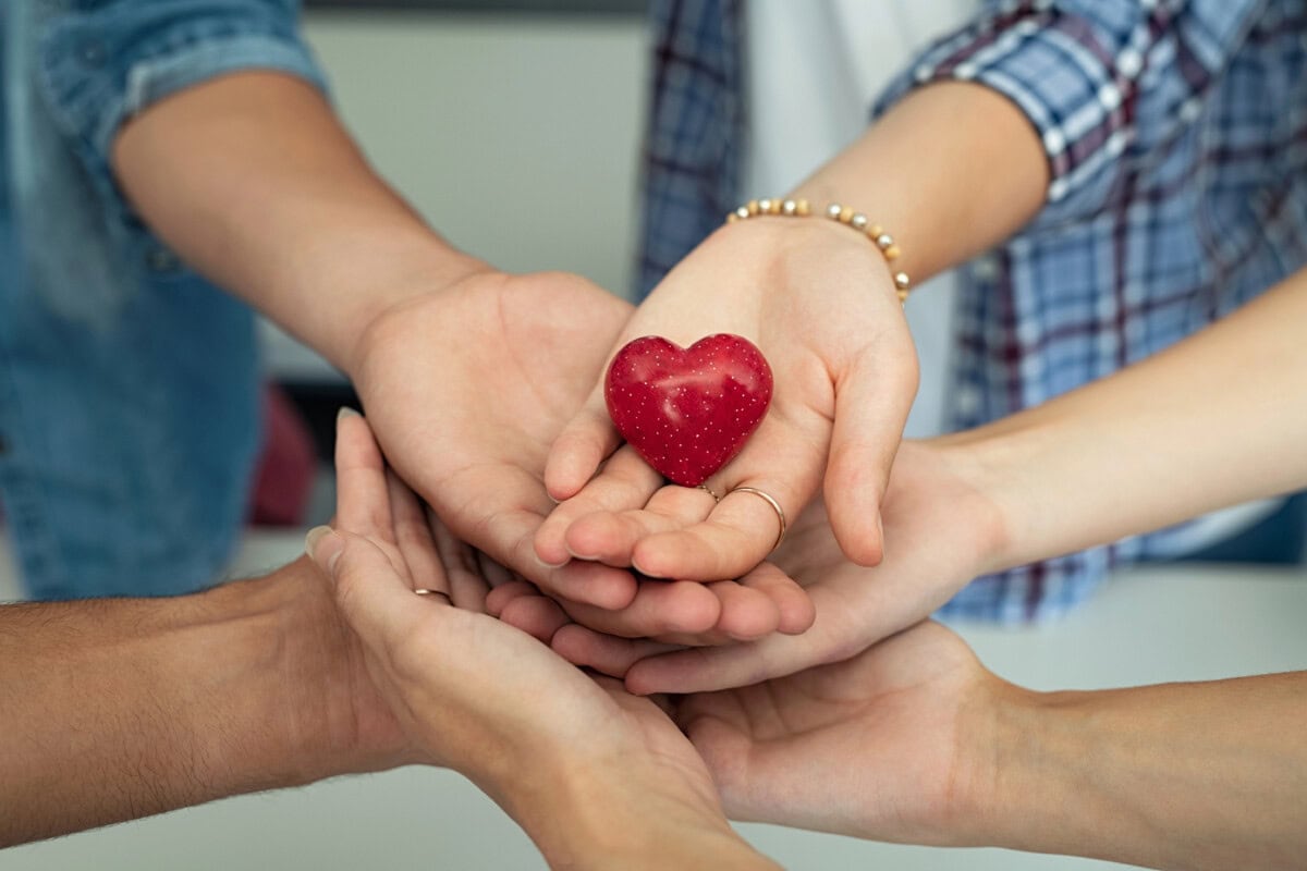 Several hands are cupped together, holding a small red heart-shaped object.