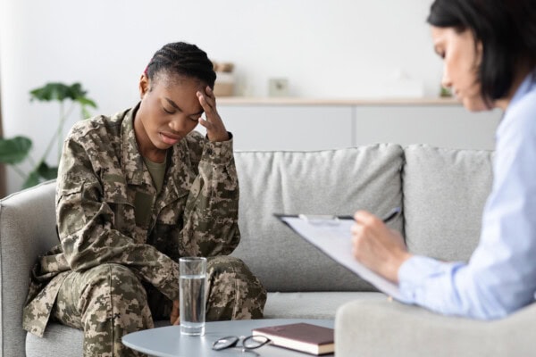 A woman in military uniform sits on a couch with her head in her hand, while another woman takes notes on a clipboard during a counseling session.