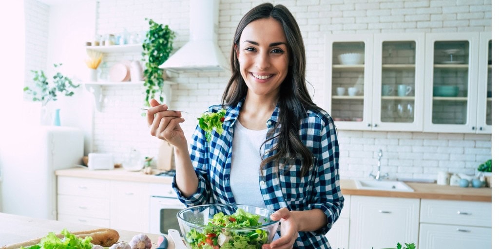 A woman stands in a bright kitchen, smiling while holding a fork and a glass bowl of salad.