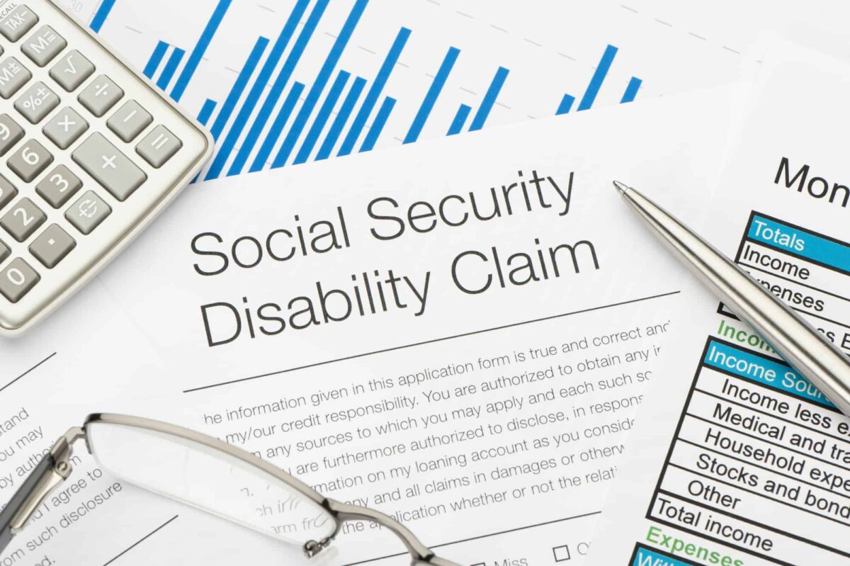 A Social Security Disability Claim form on a desk, surrounded by a calculator, eyeglasses, a pen, and financial documents with charts and tables.