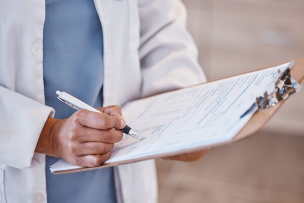 A person in a lab coat writes on a clipboard filled with documents or forms.