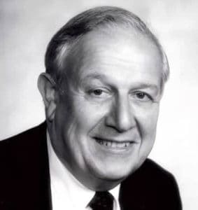 Black and white portrait of an older man with short hair, wearing a suit and tie, smiling at the camera against a plain background.