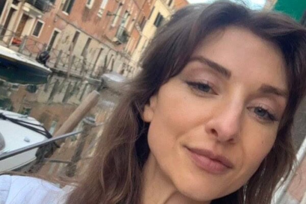 A woman takes a selfie near a canal lined with boats and brick buildings.