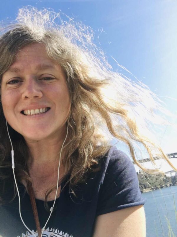 Woman with wavy blonde hair smiles at the camera outdoors, wearing a navy shirt and white earphones, with blue sky and water in the background.