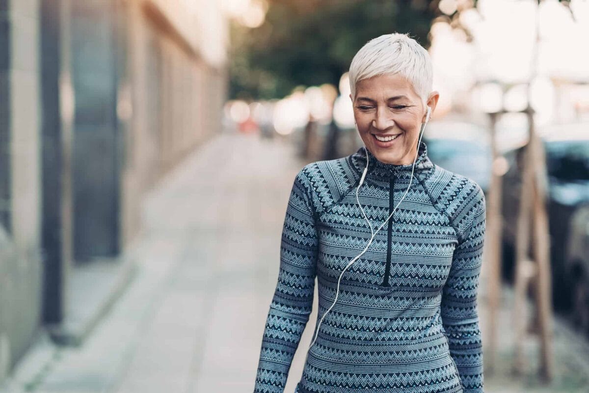 Smiling woman with short gray hair walks outdoors on a city sidewalk, wearing a patterned sweater and earphones.