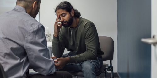 Over-the-shoulder shot of a doctor and man. The man looks sad and depressed after hearing news from the doctor. The man's eyes are closed and has one hand covering part of his face.