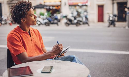 Modern you man taking notes while sitting in cafe by the street in city.