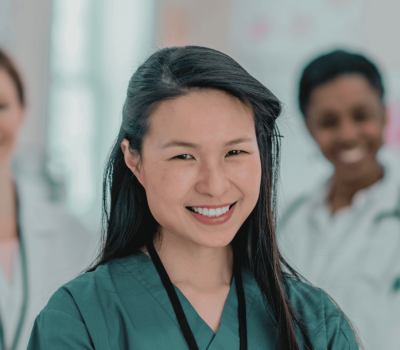 A woman in medical scrubs smiles at the camera, with four healthcare professionals standing blurred in the background.