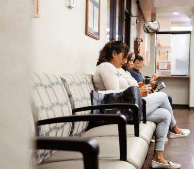Three people sit and look at their phones in a waiting room with empty chairs and a reception desk in the background.