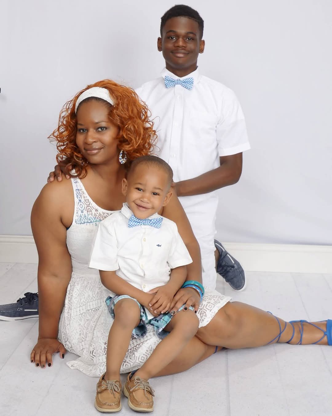 A woman sits on the floor with a young boy on her lap and an older boy kneeling beside them. All are dressed in white outfits and blue accessories against a plain background.