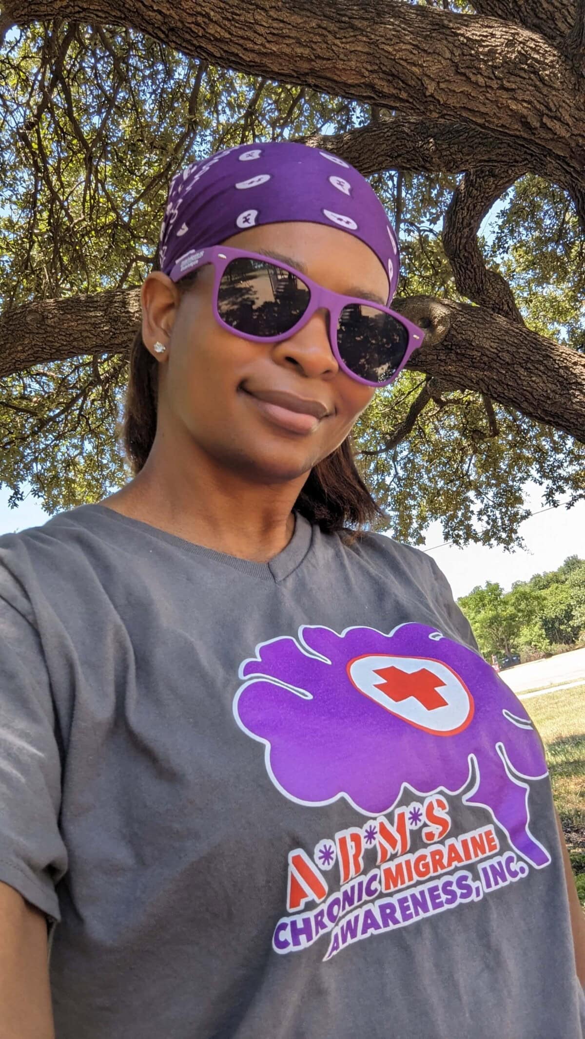 A woman wearing purple sunglasses and a bandana smiles outdoors. Her shirt has a brain graphic and reads, "Chronic Migraine Awareness, Inc." Tree branches are visible in the background.