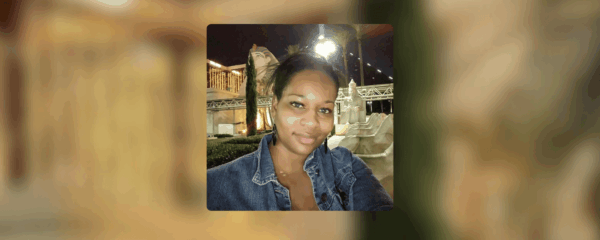 A woman in a denim jacket takes a selfie outdoors at night, with illuminated structures and statues visible in the blurred background.