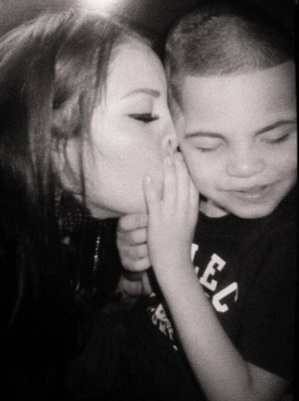 A woman kisses a young boy on the cheek while he smiles, both appearing happy in a black and white photo.
