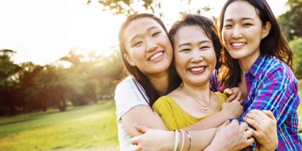 Three women standing outdoors in a park, smiling and embracing each other, with sunlight in the background.