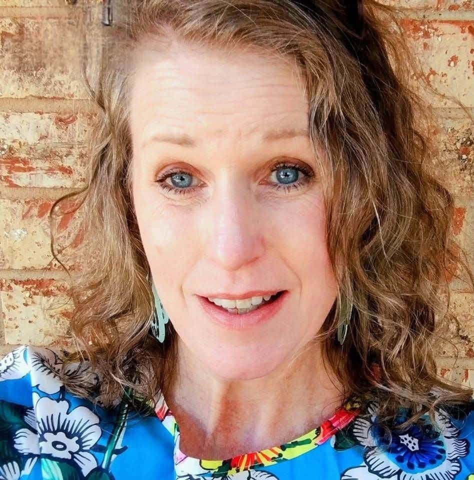 Woman with curly light brown hair and blue eyes, wearing a blue floral top, poses in front of a brick wall, looking directly at the camera.