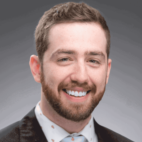 A man with short brown hair and a beard, wearing a dark suit, white shirt with a pattern, and a light blue tie, smiles at the camera against a gray background.