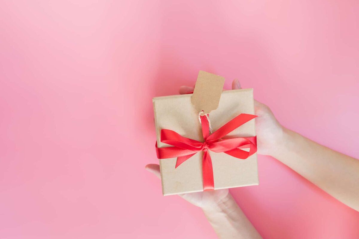 A person holds a gift wrapped in brown paper with a red ribbon and a blank tag against a pink background.