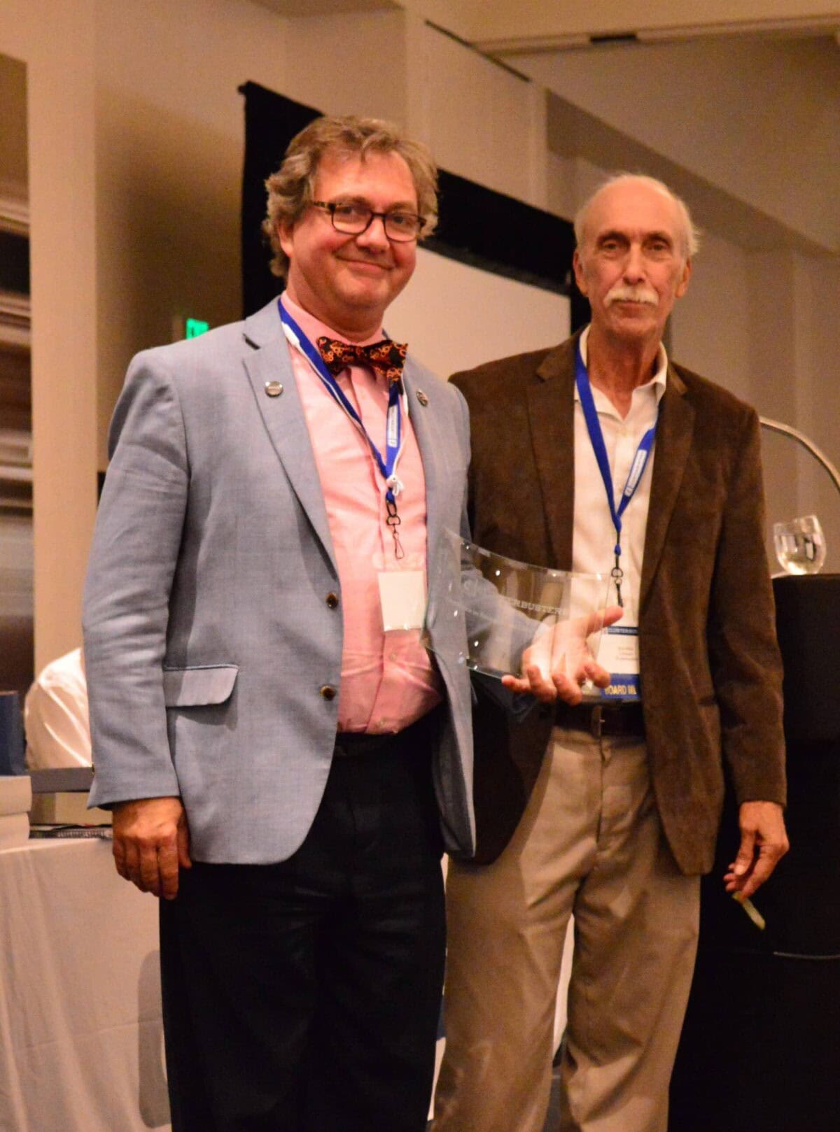 Two men stand side by side at an indoor event; one holds a clear glass award, both wear jackets and event lanyards.