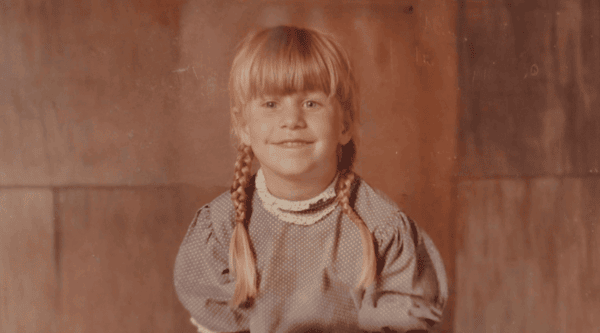 A young child with light hair in two braids smiles at the camera, wearing a polka dot dress with a lace collar, posed against a wooden backdrop.