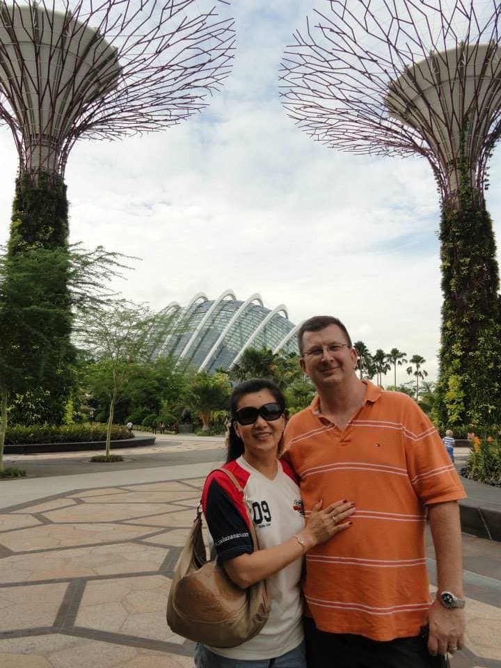 A man and woman pose together outdoors at Gardens by the Bay in Singapore, with Supertree structures and the Flower Dome visible in the background.