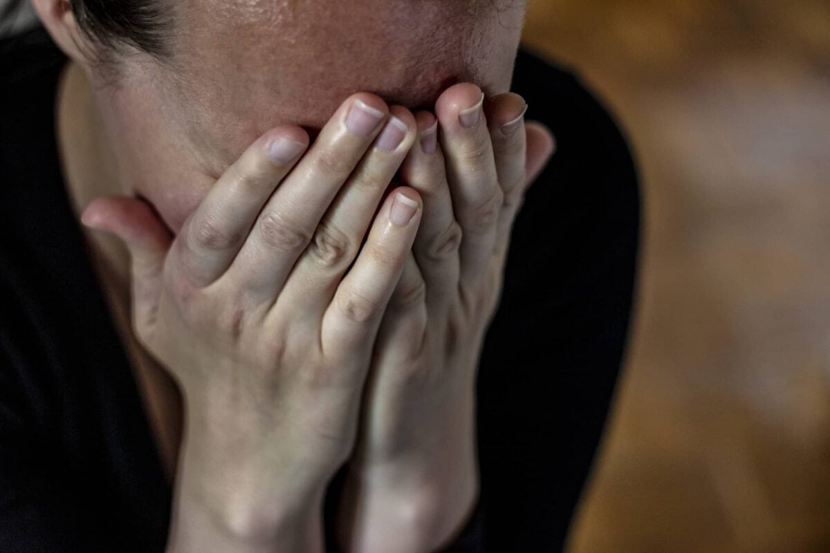 A person with dark hair covers their face with both hands, obscuring their expression. The background is out of focus.