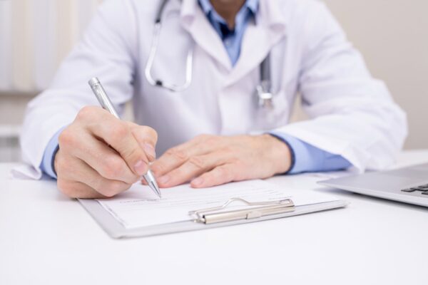 A doctor wearing a white coat and stethoscope writes on a clipboard with a pen at a desk.
