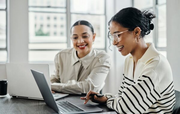 Two women are sitting at a desk with open laptops, one smiling and the other typing, in a bright office setting.