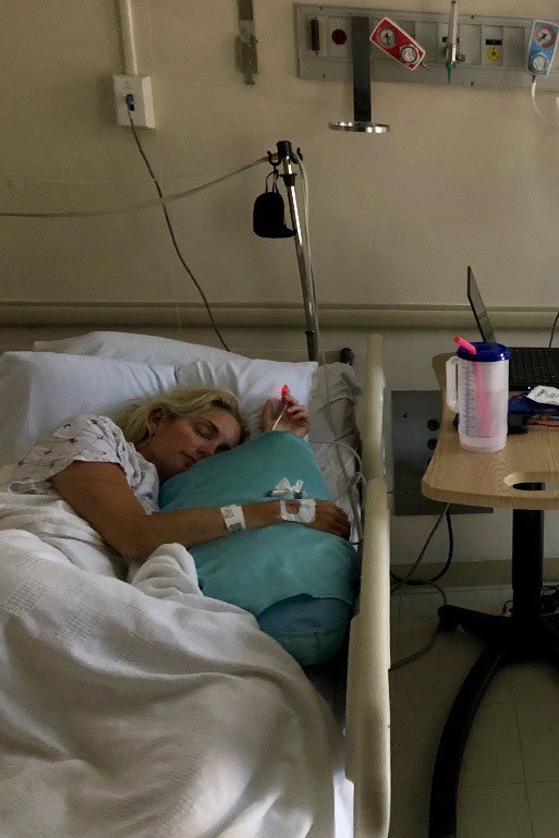 A woman in a hospital bed, wearing a hospital gown, hugs a blue pillow while lying on her side. Medical equipment and a water bottle are visible beside her.
