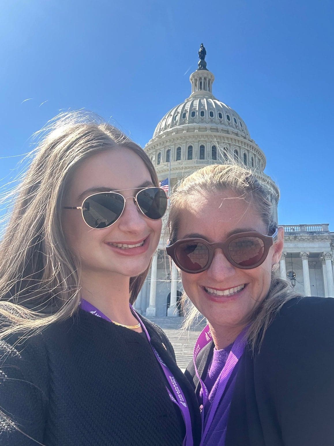 Two women wearing sunglasses smile in front of the U.S. Capitol building on a sunny day.
