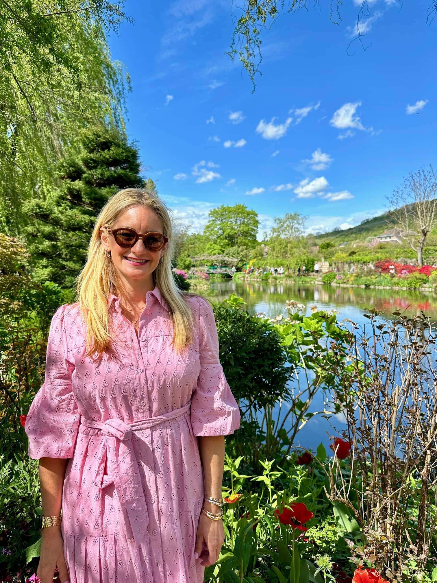 A woman in a pink dress and sunglasses stands in a garden beside a pond, with trees, flowers, and a blue sky in the background.