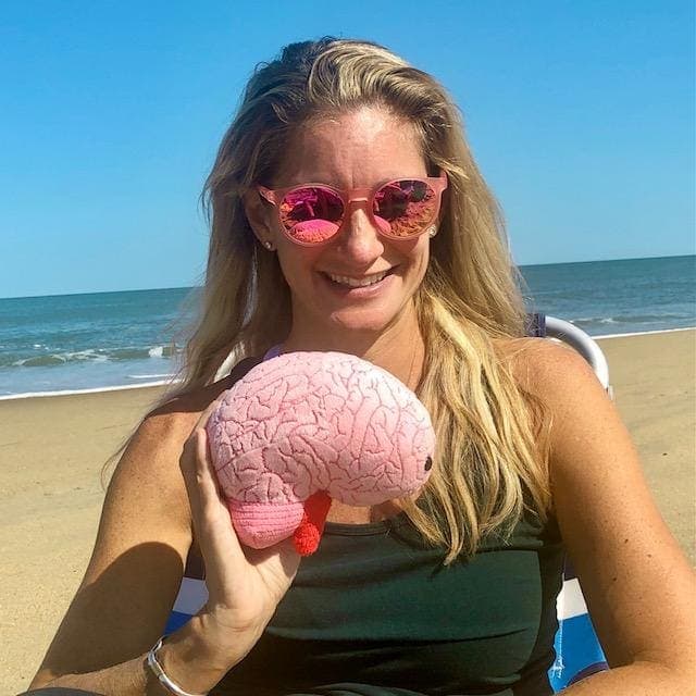 A woman sitting on a beach holds a pink plush toy shaped like a brain, wearing pink sunglasses and smiling at the camera.