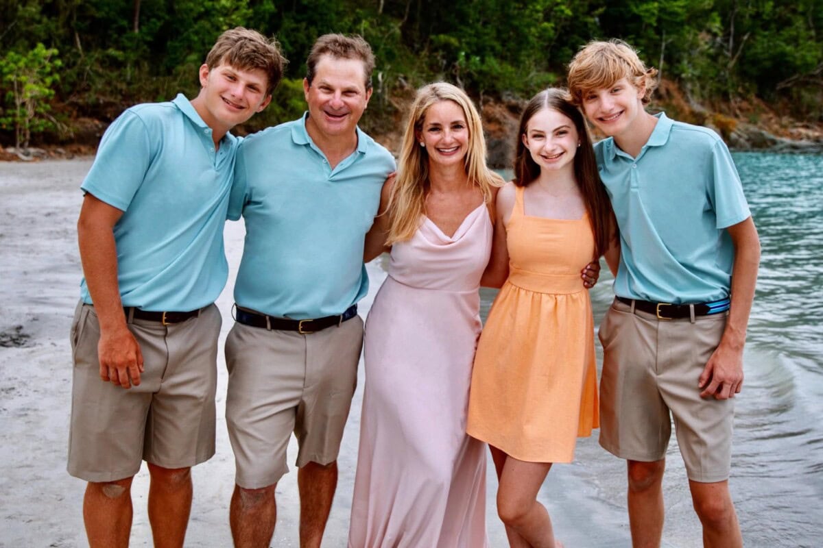 A family of five poses on a beach, with three males in matching light blue shirts and khaki shorts, and two females in pastel dresses, smiling in front of trees and water.