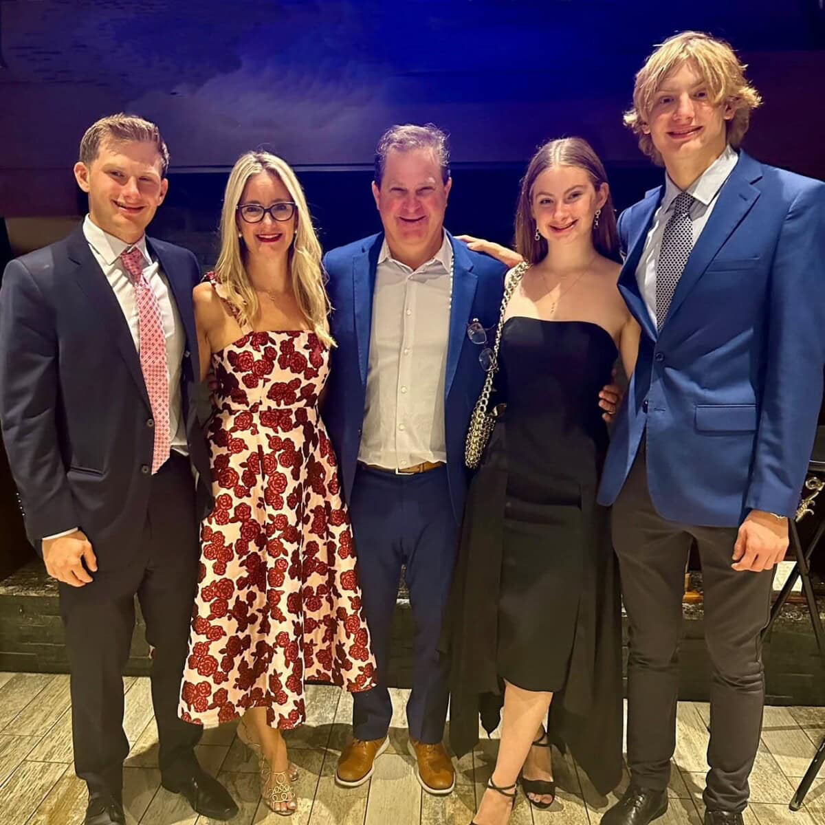 Five people dressed in formal attire stand closely together indoors, smiling at the camera against a dark background.