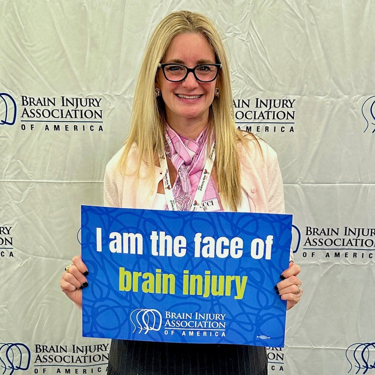 A woman stands in front of a Brain Injury Association of America backdrop, holding a sign that reads, "I am the face of brain injury.