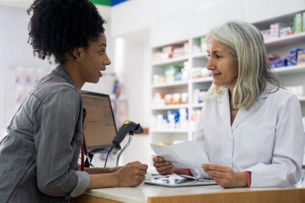 A pharmacist in a white coat speaks with a customer at the pharmacy counter, holding a piece of paper and standing in front of shelves stocked with products.