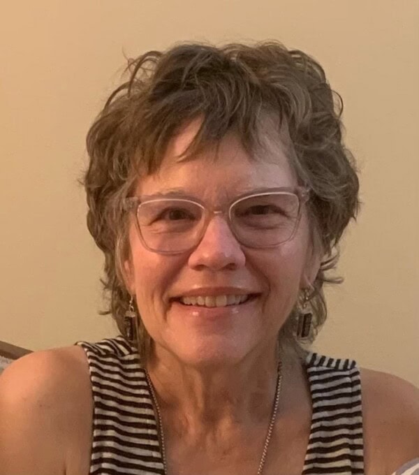 A woman with short, curly hair and glasses smiles at the camera. She is wearing a sleeveless, striped top and a necklace. The background is a plain beige wall.