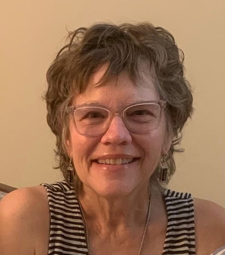 A woman with short, curly hair and glasses smiles at the camera. She is wearing a sleeveless, striped top and a necklace. The background is a plain beige wall.
